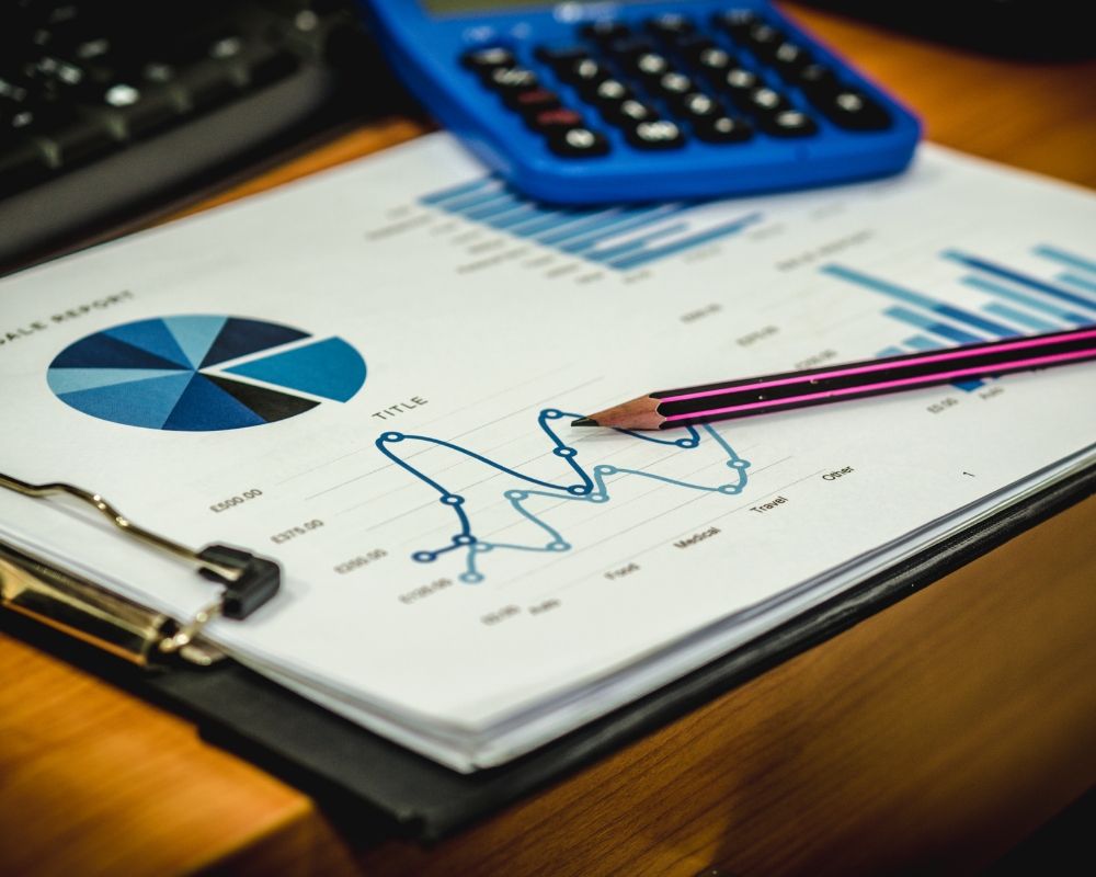 A decorative image showing many charts on a piece of paper that's attached to a clipboard. There is a pen, calculator and the edge of a keyboard in the picture.